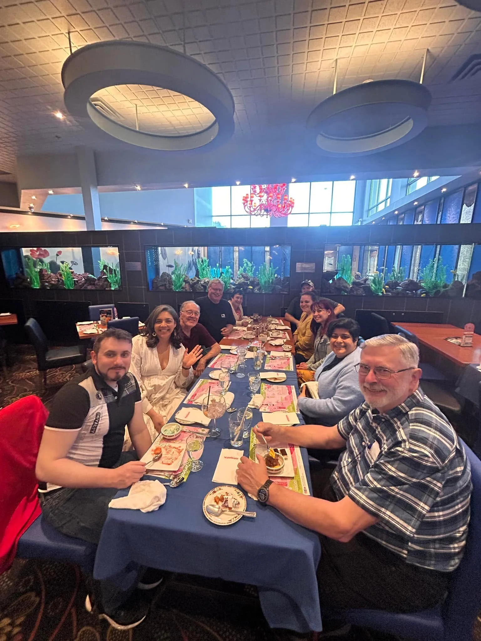 A group of people sitting at a long table in a restaurant, smiling and posing for a photo. The table has pink menus, glasses, and dishes. The restaurant has large windows, a red chandelier, and fish tanks on the wall.