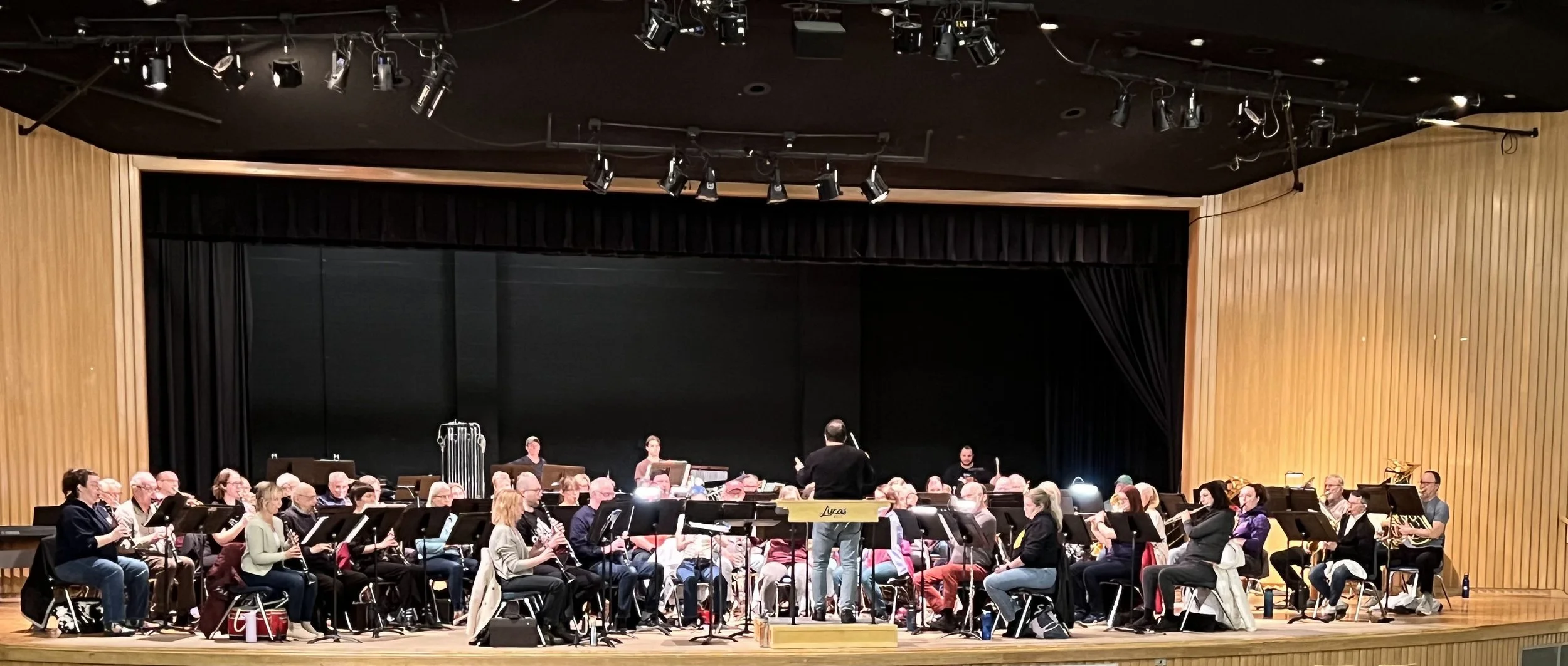 A large group of musicians sitting on stage with a conductor standing in front of them, practicing or performing in a concert hall with wooden walls and stage lighting.