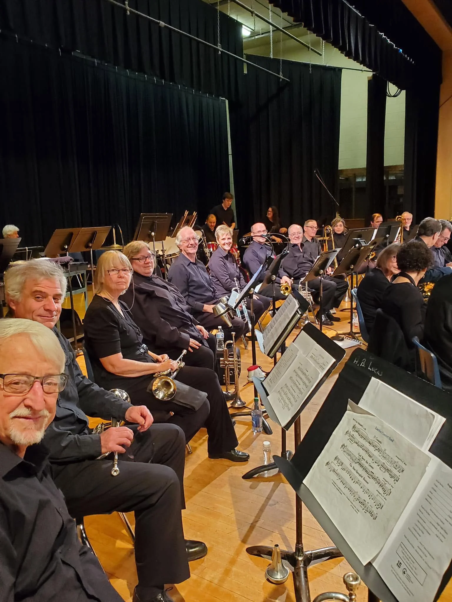 An orchestra seated on stage with sheet music stands in front of them. Musicians hold various instruments and are dressed in formal black attire. A black curtain backdrop and stage equipment are visible.