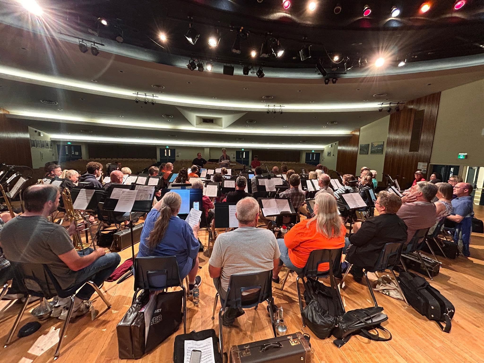 Orchestra rehearsal in a large concert hall, with musicians seated and practicing their instruments while a conductor leads the group. The room has bright overhead lighting and a wooden floor.