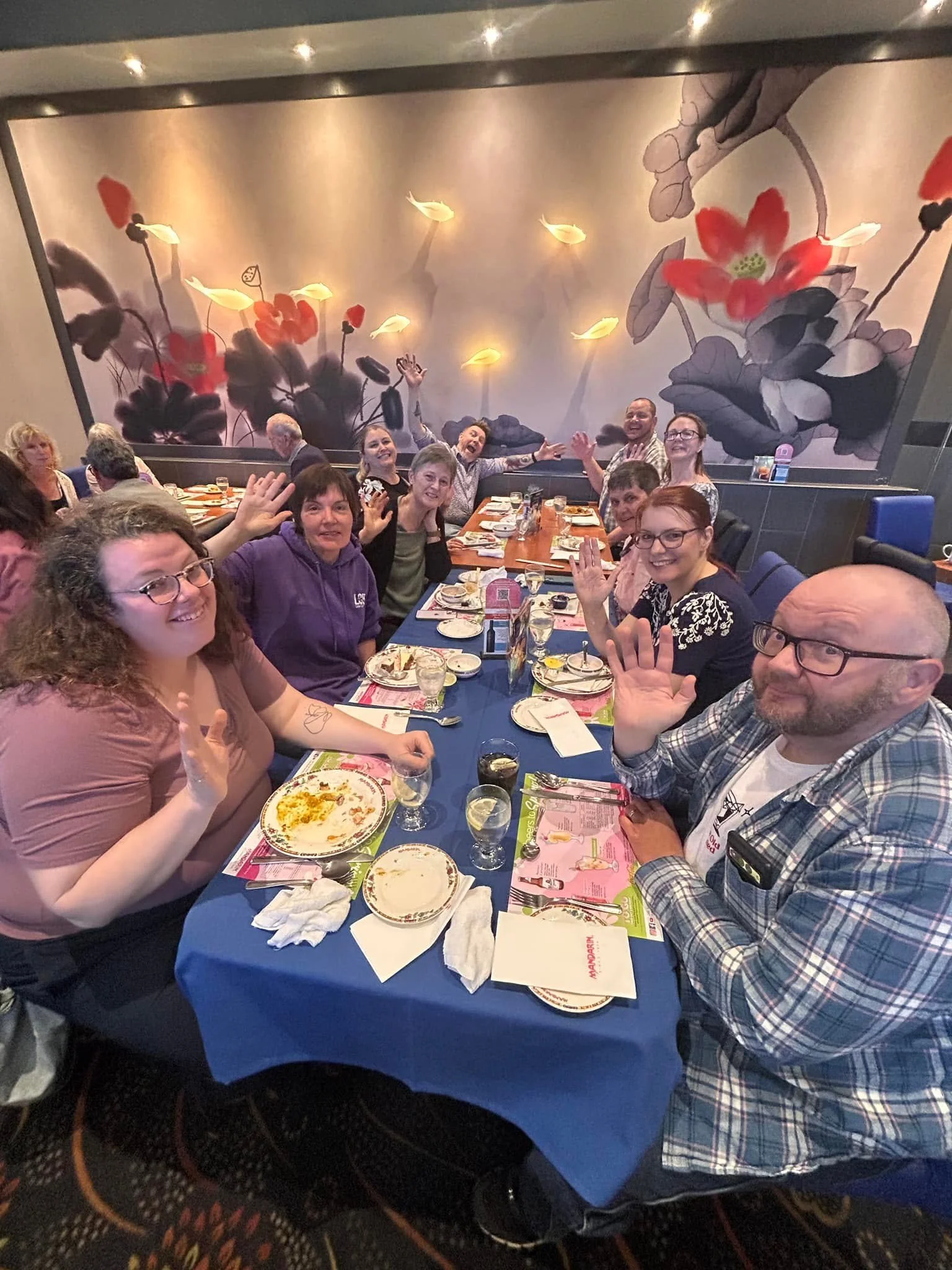 People gathered around a table at a restaurant, smiling and waving, with a large decorative mural of flowers on the wall behind them.