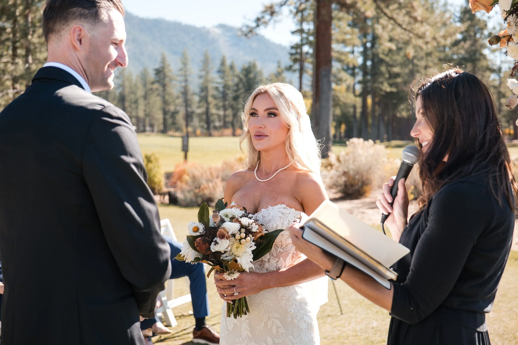An Edgewood bride with blonde hair in a strapless white wedding dress holding a bouquet of flowers, standing outdoors at a wedding ceremony in South Lake Tahoe. Officiant from Pomegranate Occasions.