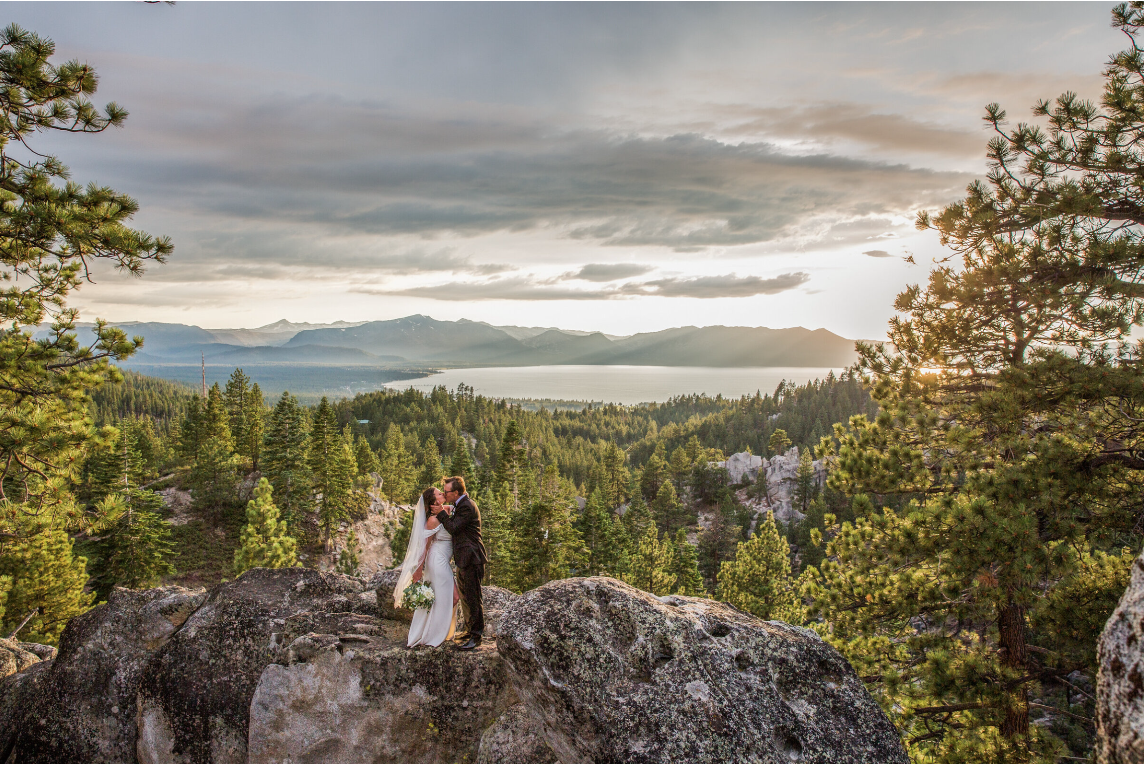 A bride and groom embracing outdoors, with the groom in a black tuxedo and the bride in a white wedding gown and veil, standing near bushes with a body of water and cloudy sky in the background.