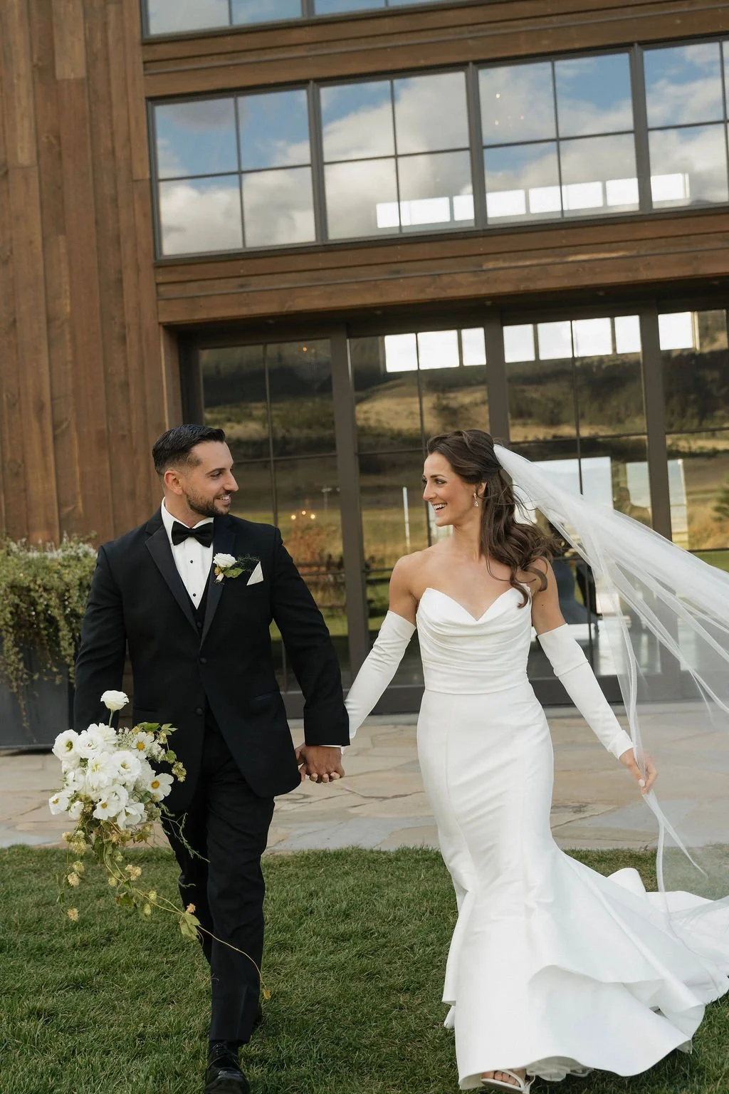 groom holding bouquet while bride and groom look at each other and walk away from the barn toward the camera