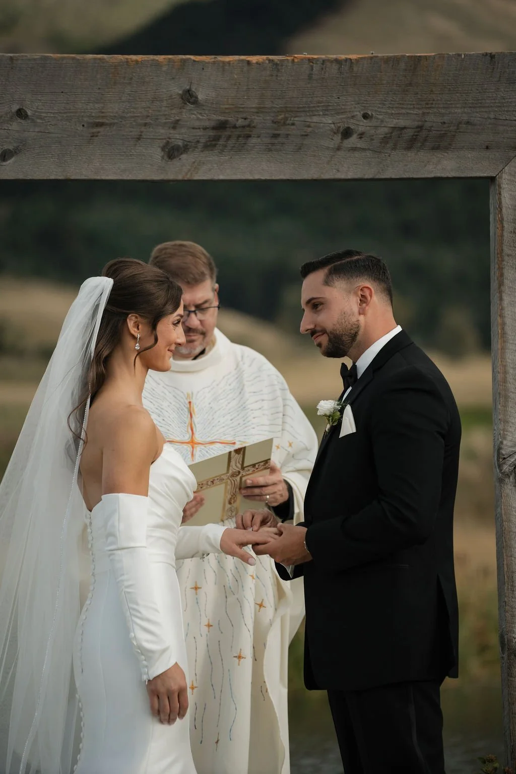 the groom giving the bride her ring with her satin dress and veil and he's wearing a black suit with the catholic priest behind