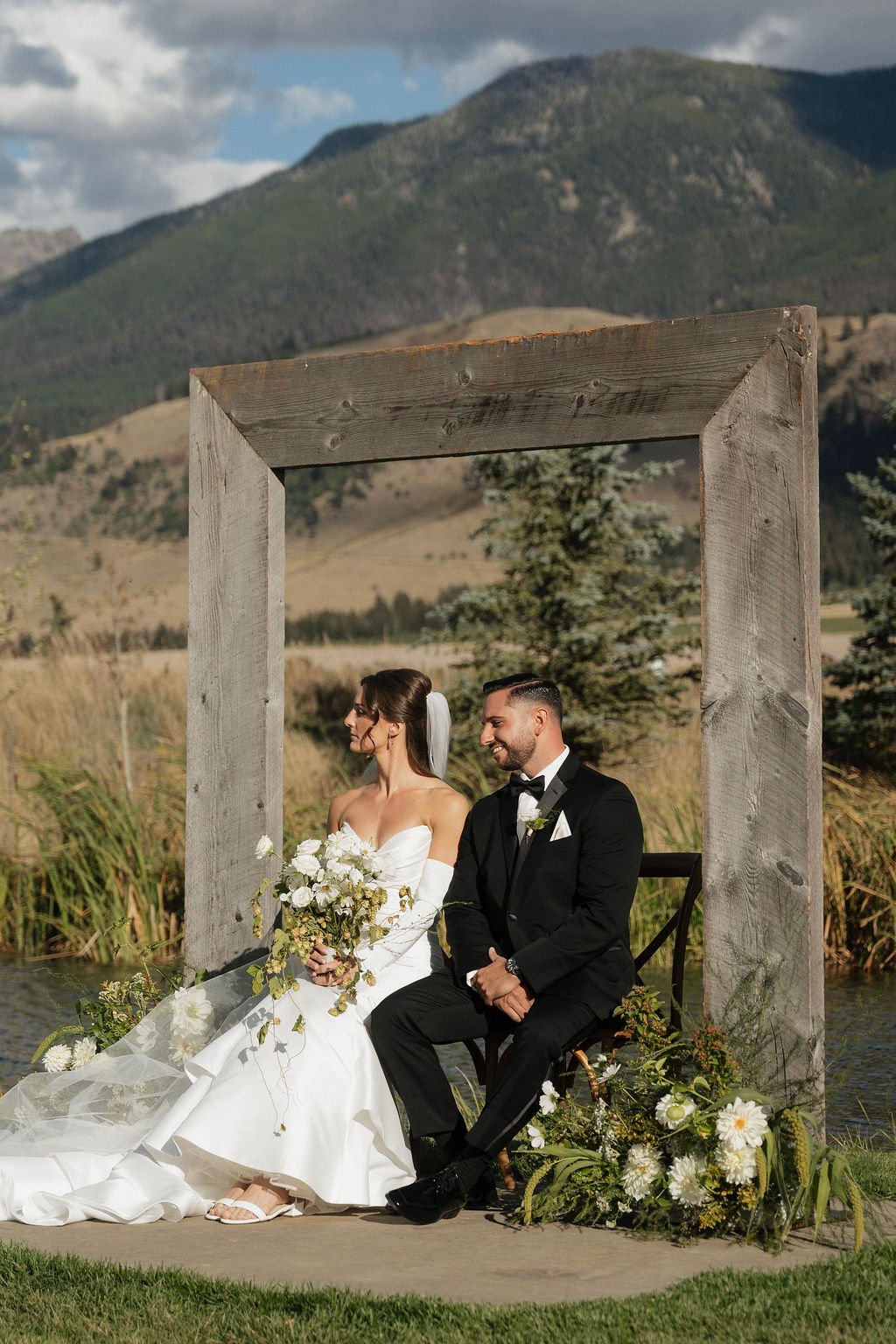 a different angle of the bride and groom sitting in the arch way with the flowers beside them