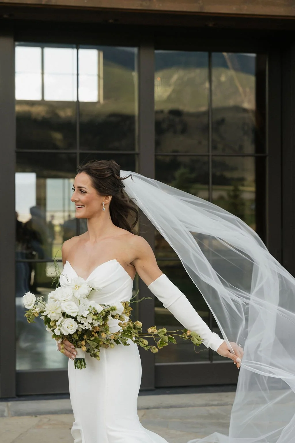 smiling bride with white wedding sleeves playing with her veil in the wind in front of the barn with a mermaid dress with her seasonal, organic garden inspired bouquet with white blooms