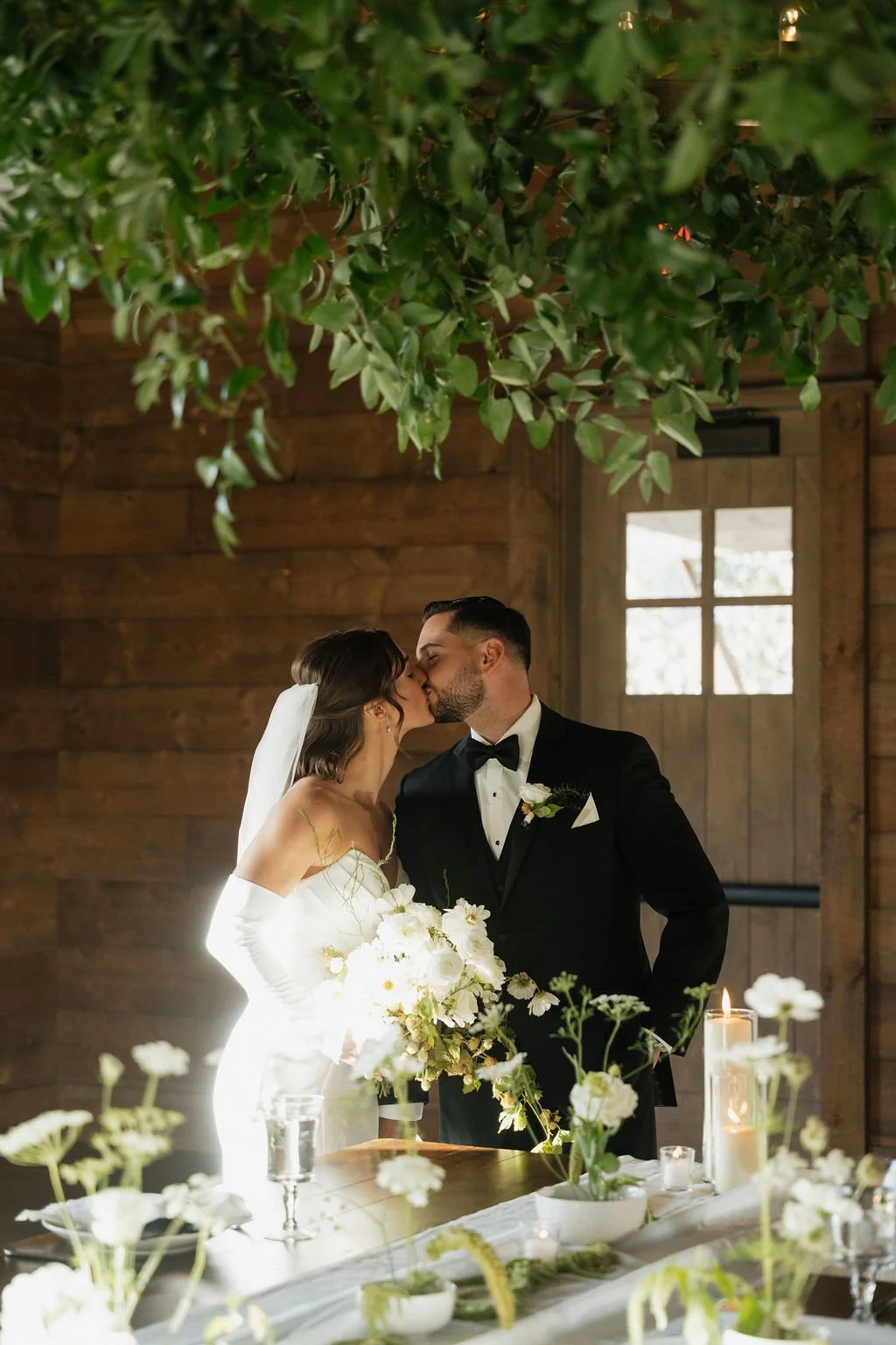 the bride and groom kissing in the reception area for the first time in warm sunshine with the chandelier framing the upper part of the picture and the ikebana arrangements on the table in front of them