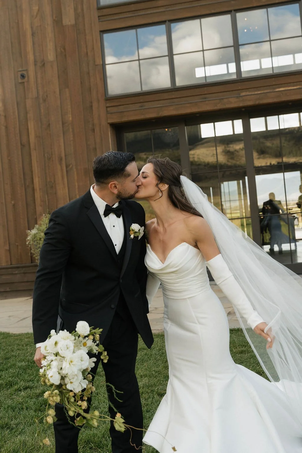 groom holding bouquet while bride and groom kiss and walk away from the barn toward the camera with her ruffly mermaid dress and his classic black tux