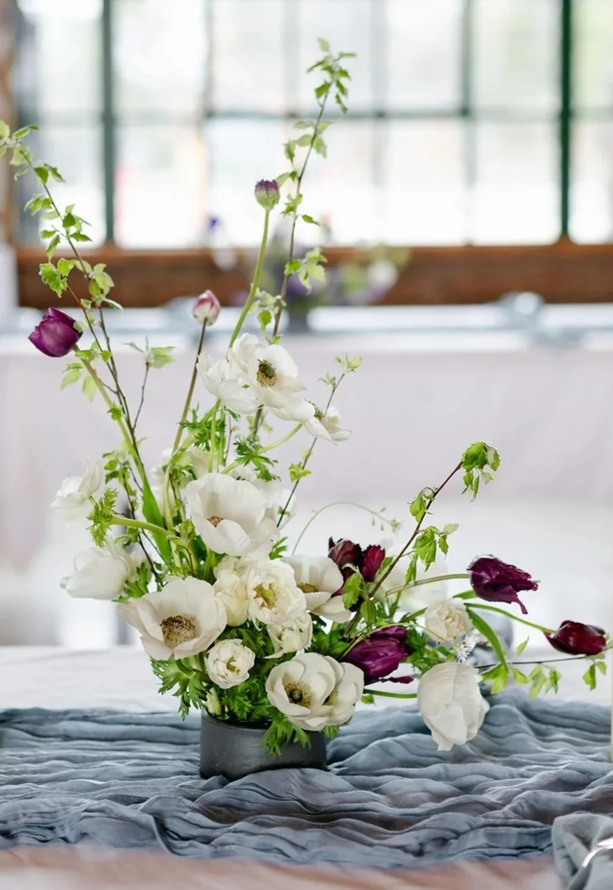 White anemones with burgundy tulips in a hand thrown vessel on a blue runner