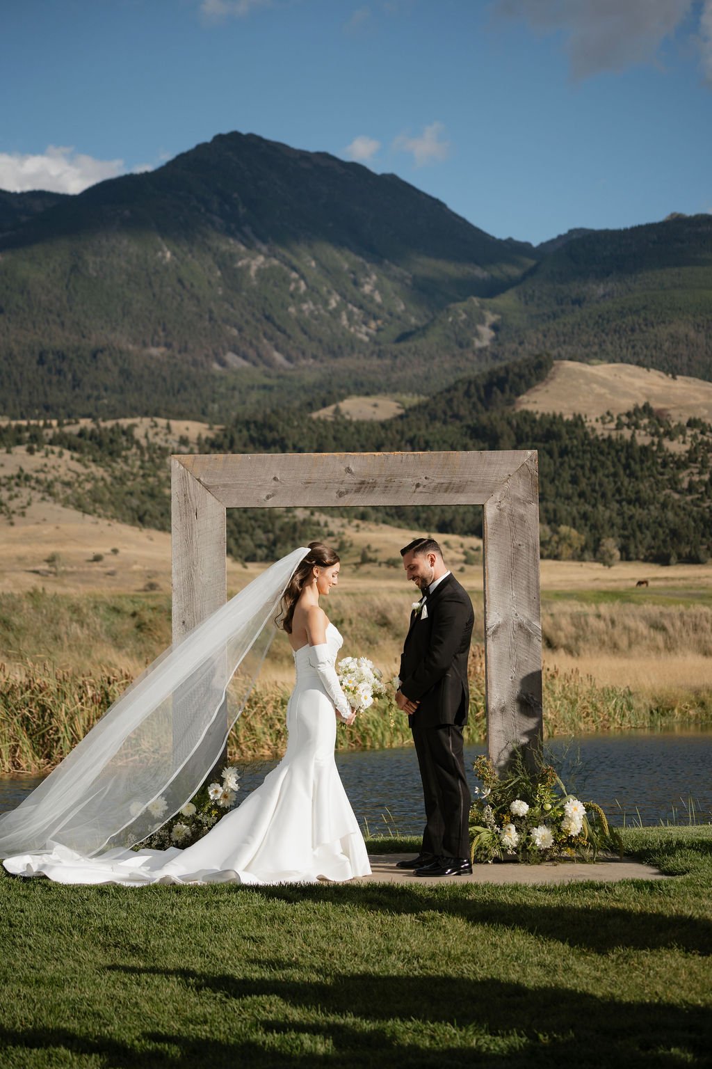 a wide angle shot of the bride and groom in front of the arch at copper rose ranch with two floral arrangements at the base with her satin mermaid dress, detached sleeves and her veil blowing in the wind