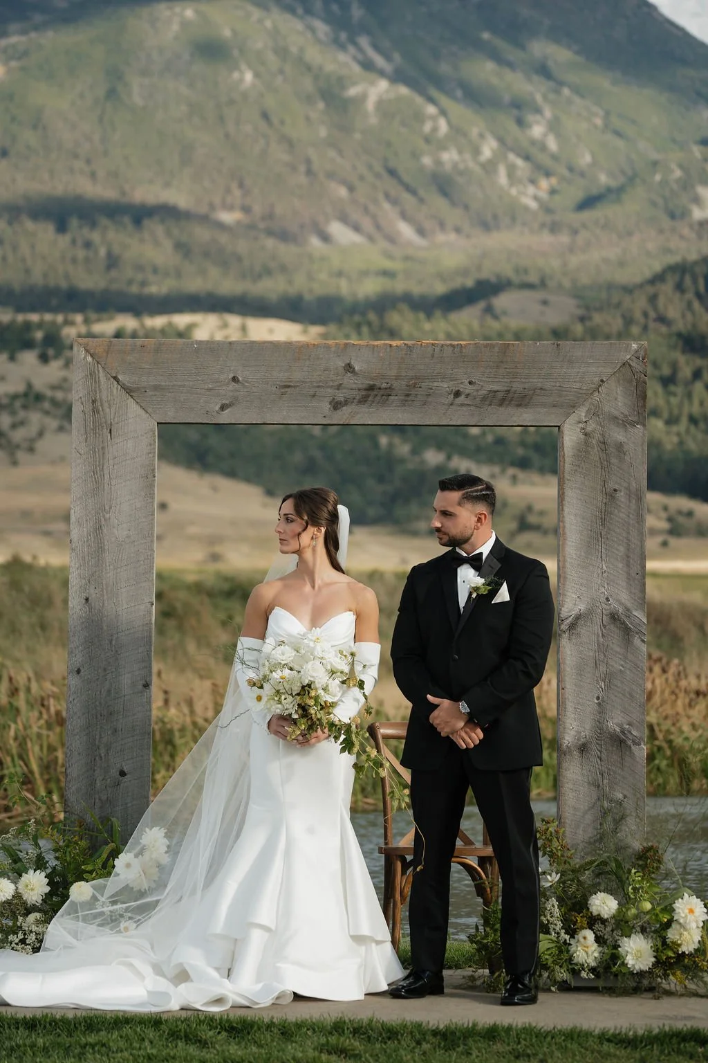 bride and groom standing listening to the catholic ceremony with the bride holding her bouquet and the groom in a black suit with the large rustic wood arch behind