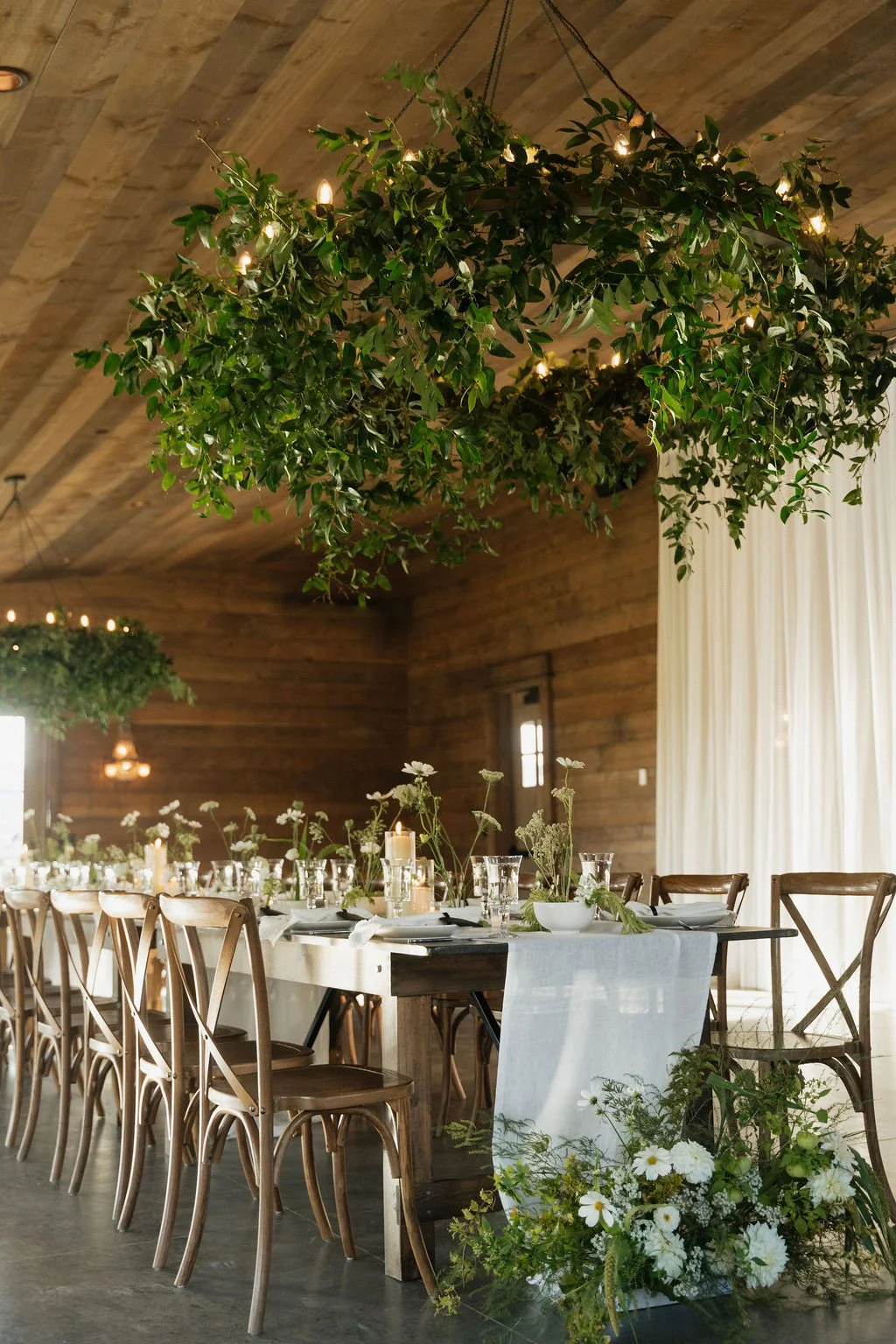 the ikebana table settings with the candles lit on farmhouse tables with black bag tags for place cards and the greenery chandelier above and one of the ceremony arrangements at the foot of the table