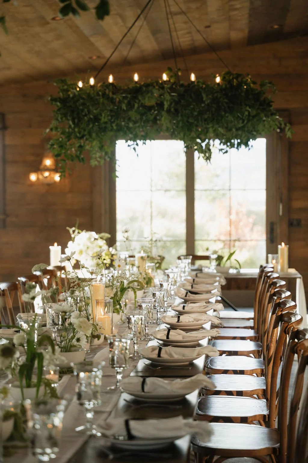 the ikebana table settings with the candles lit on farmhouse tables with black bag tags for place cards and the greenery chandelier above