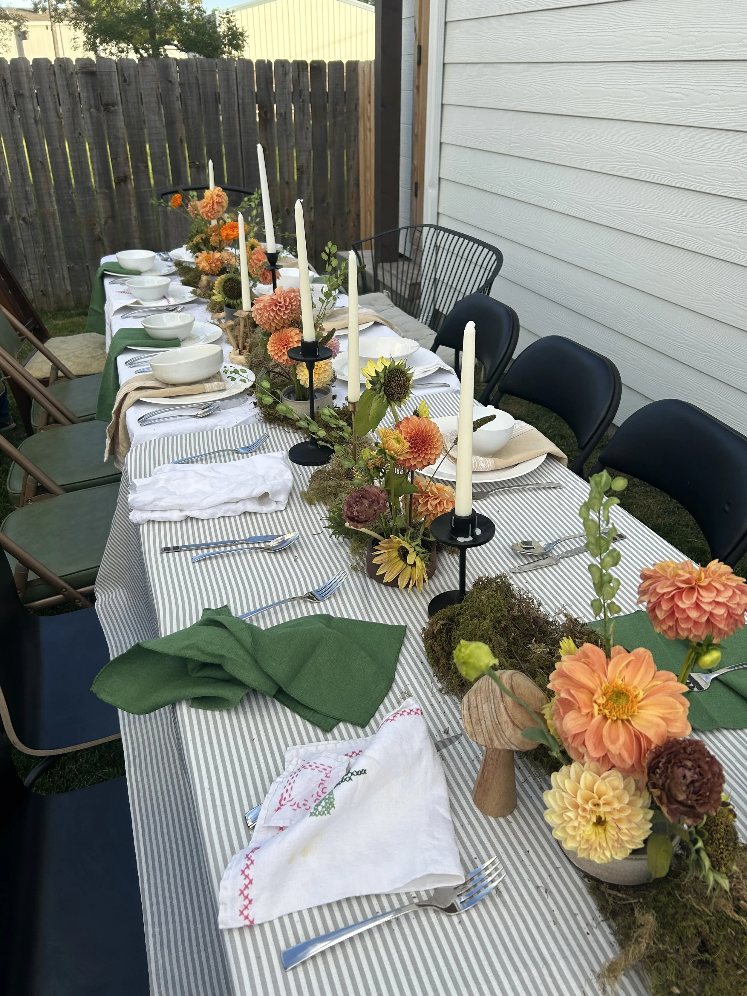 This image is of a long table with bright fall flowers on a striped linen with a jumble of chairs and a fence in the background and white taper candles in candlesticks