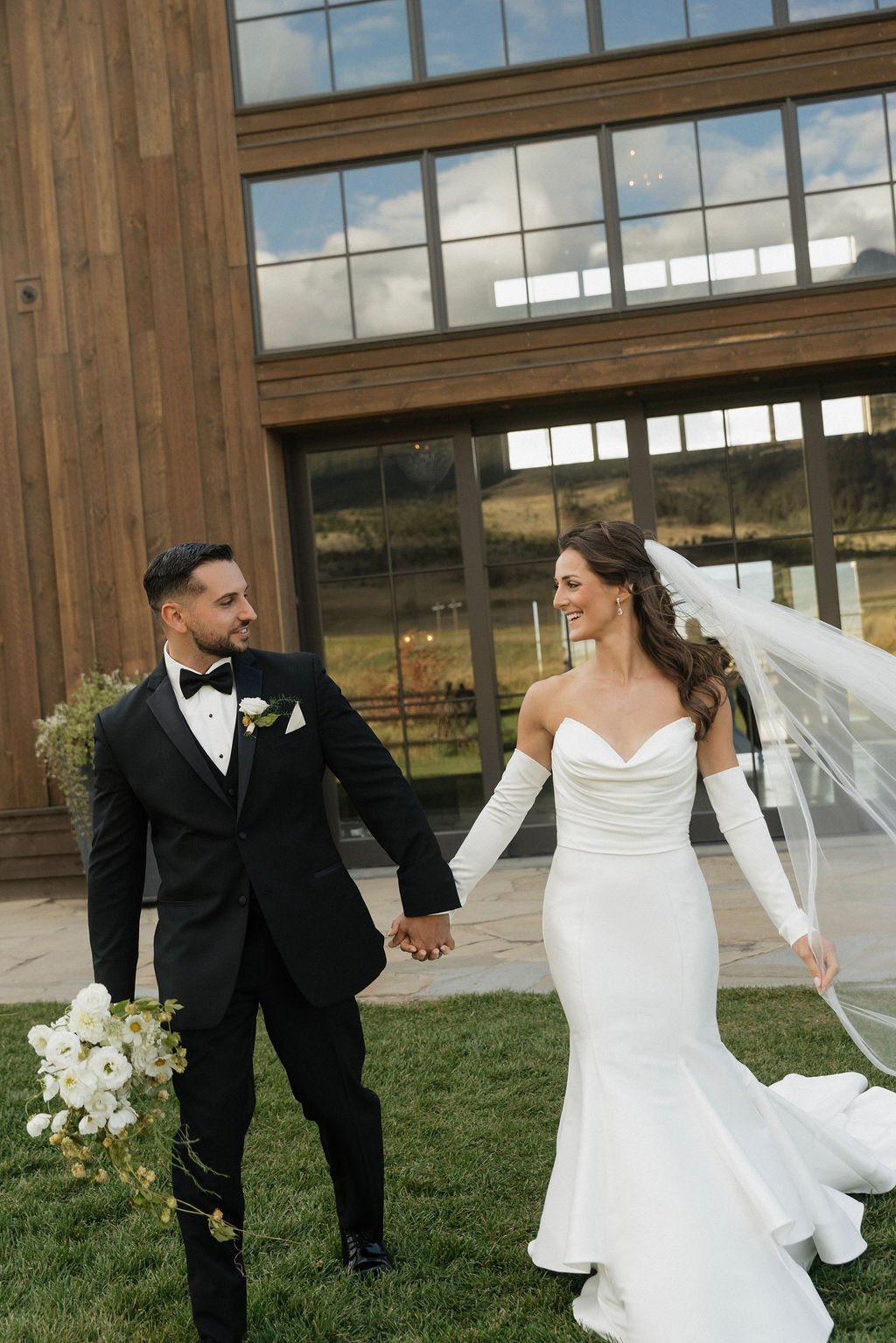 groom holding bouquet while bride and groom look at each other and walk away from the barn toward the camera with her ruffly mermaid dress 