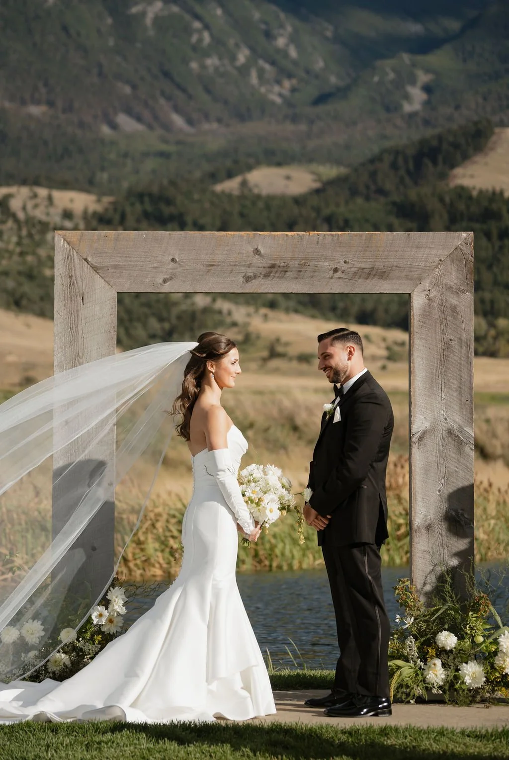 bride and groom in front of the arch at copper rose ranch with two floral arrangements at the base with her satin mermaid dress, detached sleeves and her veil blowing in the wind