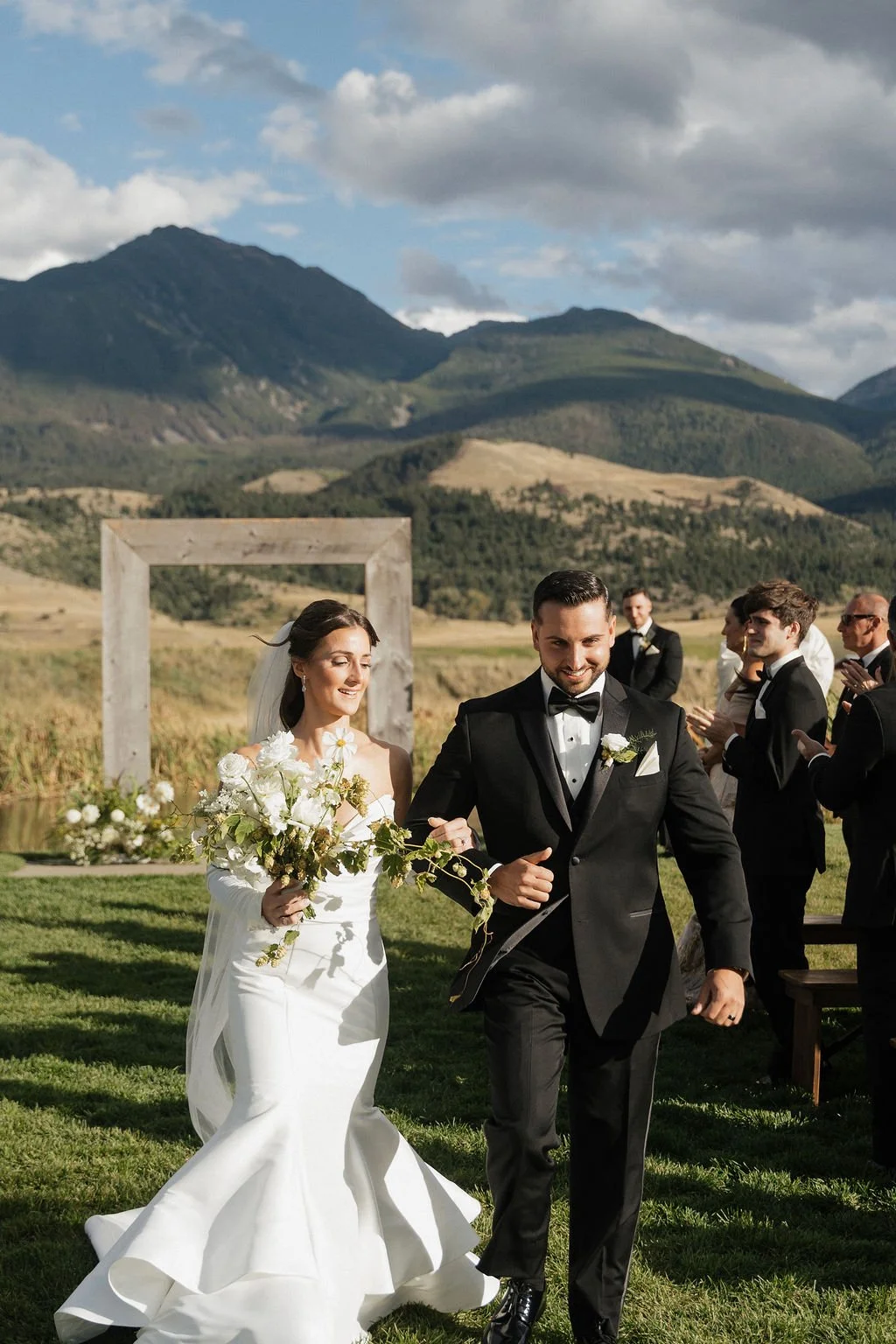 the bride and groom walking back down the aisle with big smiles on their faces the bride in a beautiful satin mermaid dress with the textural cascading bouquet in her hand