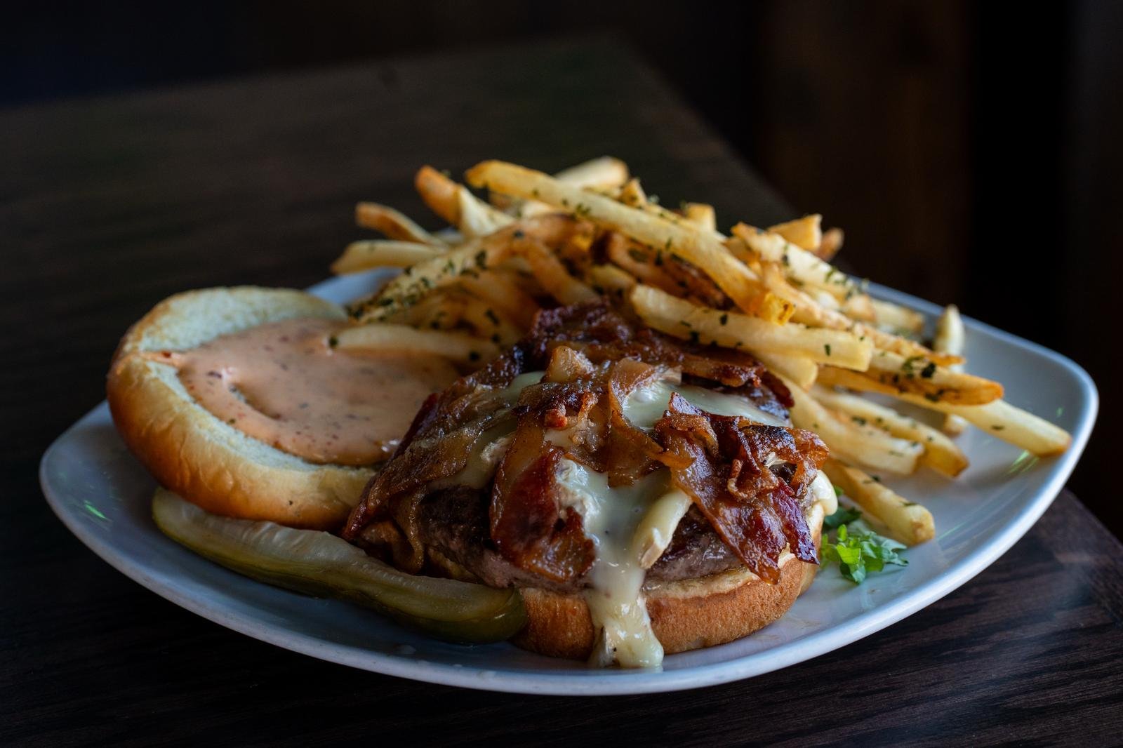 Plate of food with a burger topped with bacon and melted cheese, a side of seasoned French fries, and relish on a bun, served on a white plate on a dark wooden table.