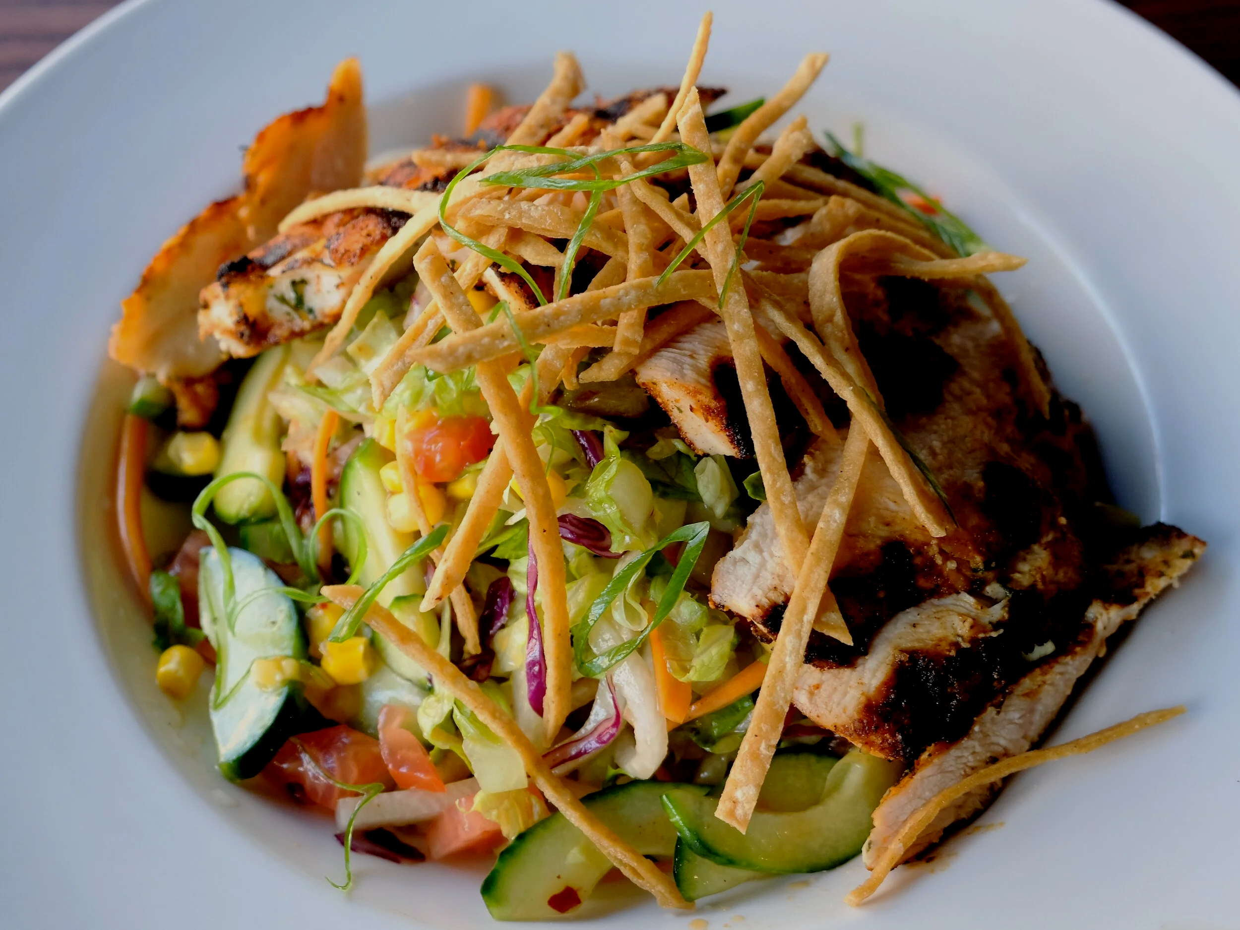 A white bowl contains a dish with grilled chicken, shredded lettuce, cucumber slices, corn, and chopped vegetables topped with crunchy tortilla strips and green onions.