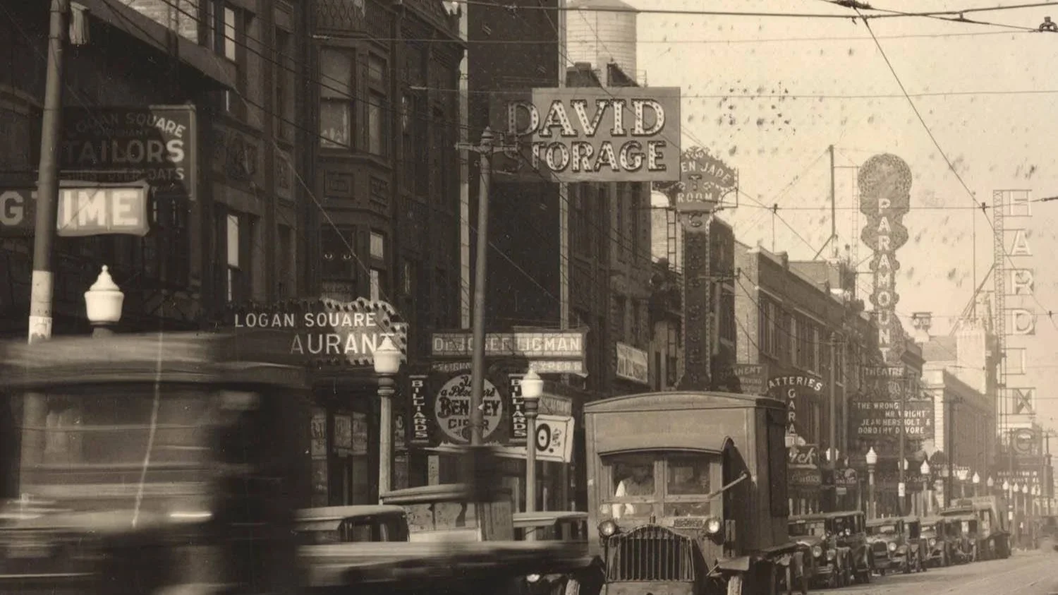 Historical city street scene with vintage trucks, signs for David Storage and Logan Square, and illuminated street lamps.