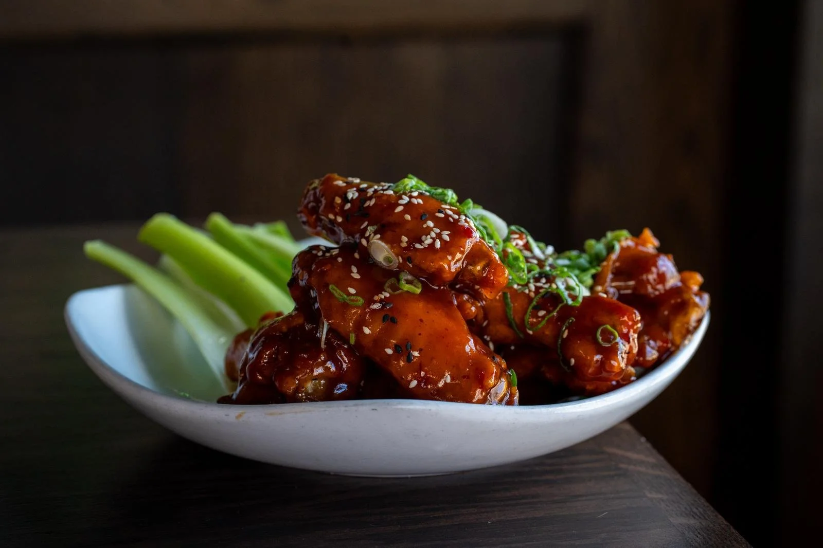 A white bowl of glazed chicken wings garnished with sesame seeds and chopped green onions, served with celery sticks on the side.