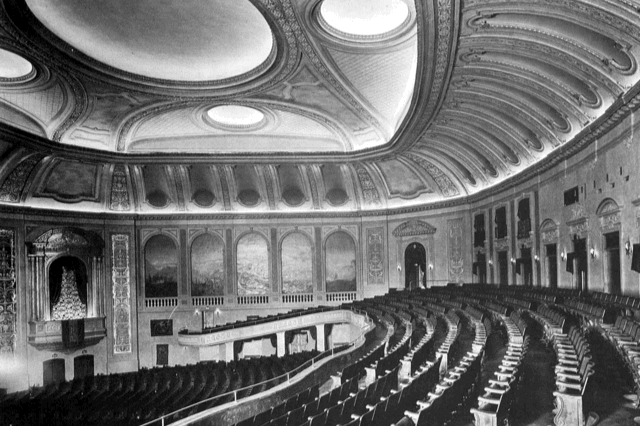 Black and white photo of an ornate theater with a curved balcony, rows of empty seats, and detailed ceiling decor.