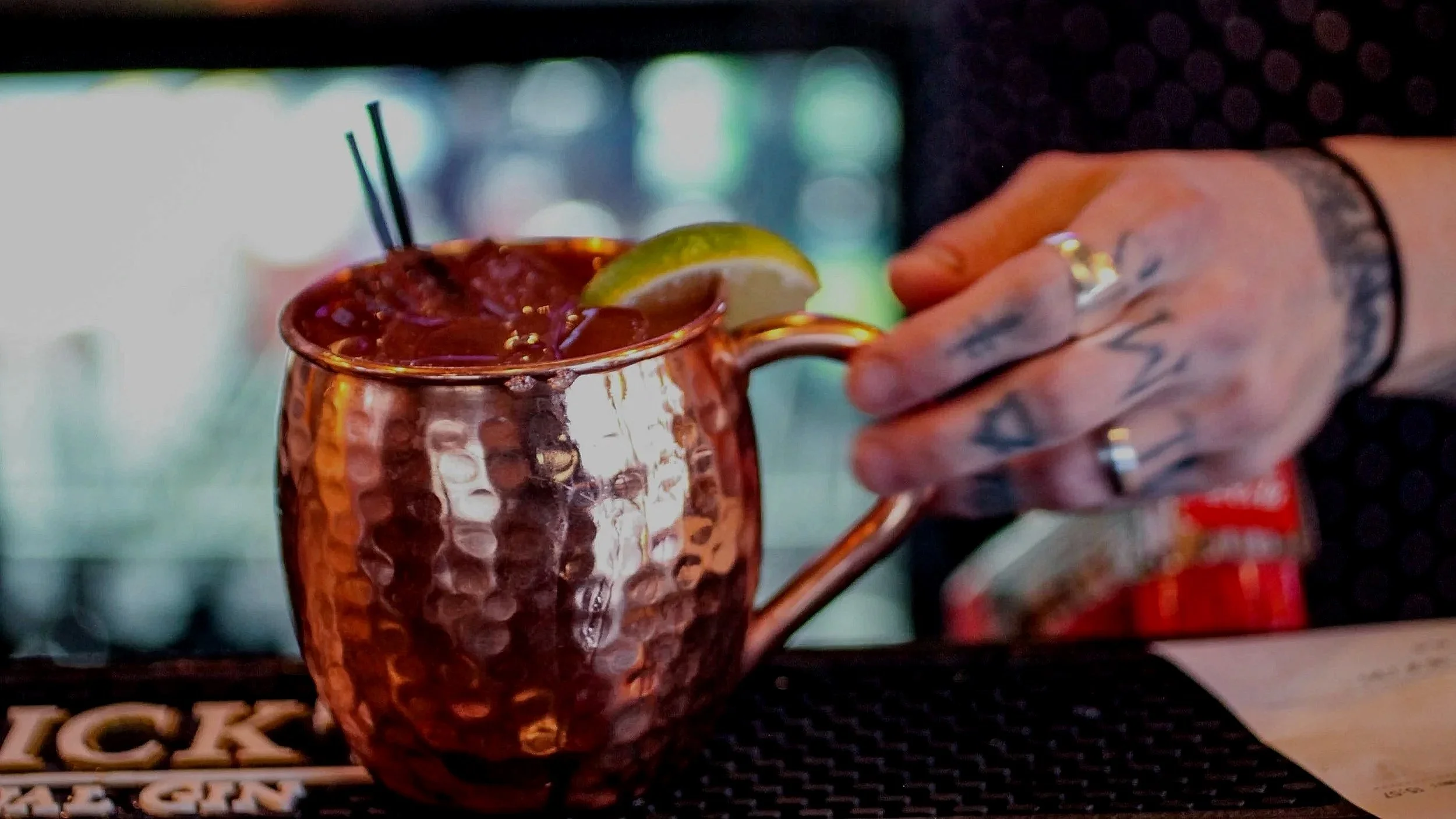 A copper mug filled with a dark beverage, garnished with a lime wedge, on a bar counter with a hand holding it.