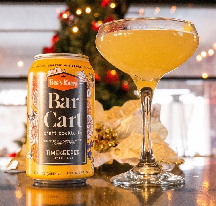 A glass full and can of Timekeeper's Bar Cart Bee's Knees cocktail sit on a bar-top with a Christmas tree in the background.