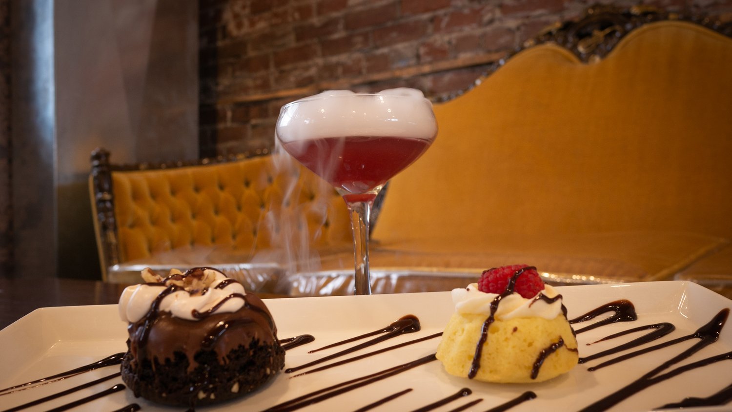 A smoking cocktail and plate of desserts sit on a coffee table next to a vintage velvet settee at Timekeeper Distillery.