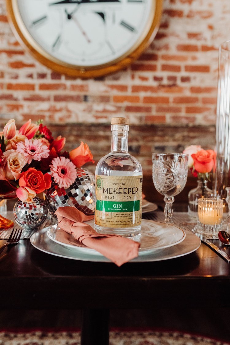A Timekeeper Distillery Gin bottle sits on the plate of a wedding place setting.