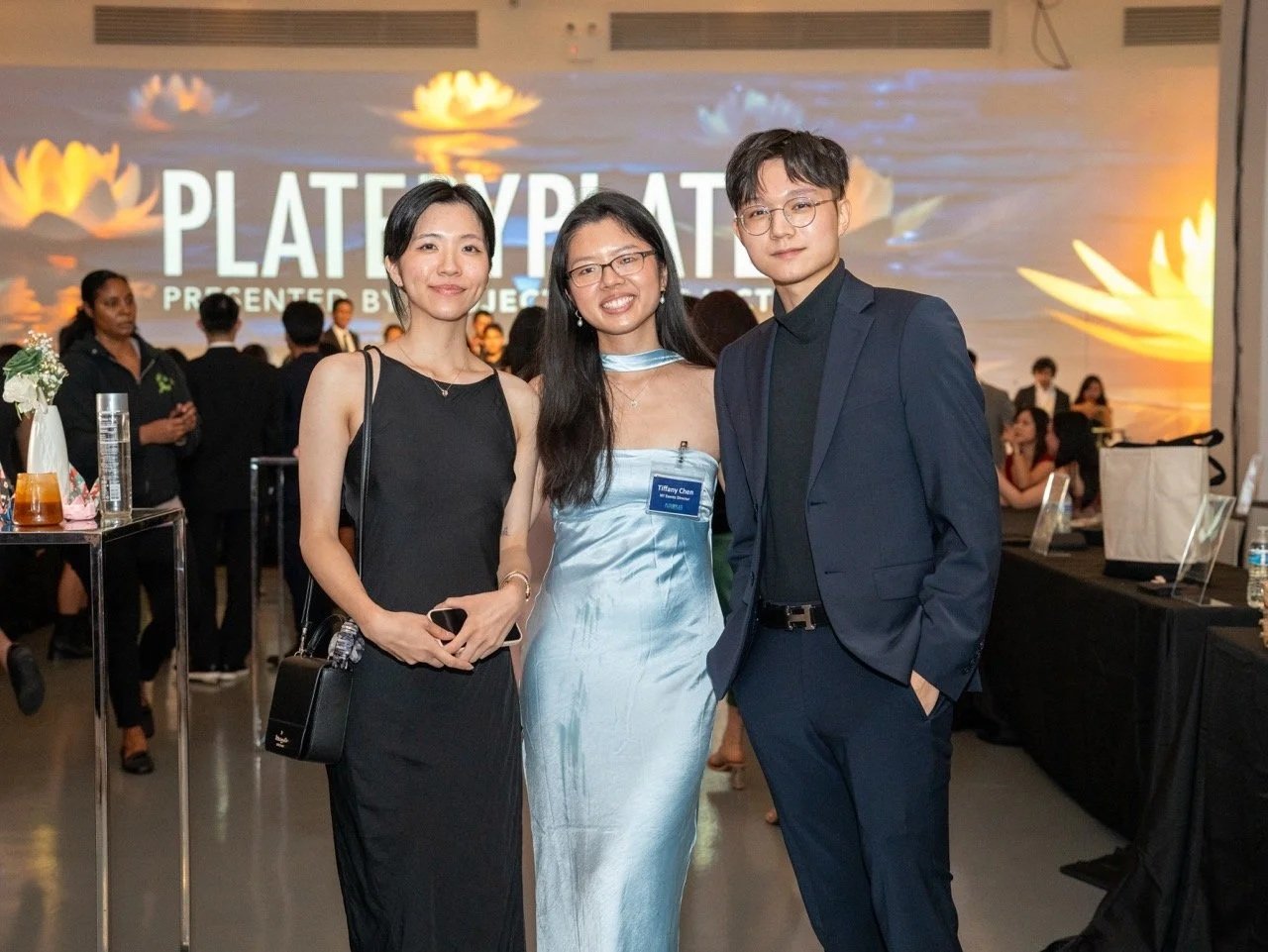 An outdoor rooftop event in a city at dusk with people socializing among trees, potted plants, and white tents, with tall buildings illuminated in the background.