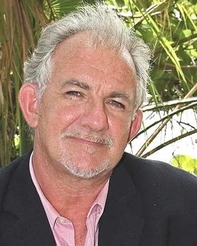 A middle-aged man with gray hair, a mustache, and a beard, wearing a pink shirt and a dark blazer, outdoors with green plants in the background.