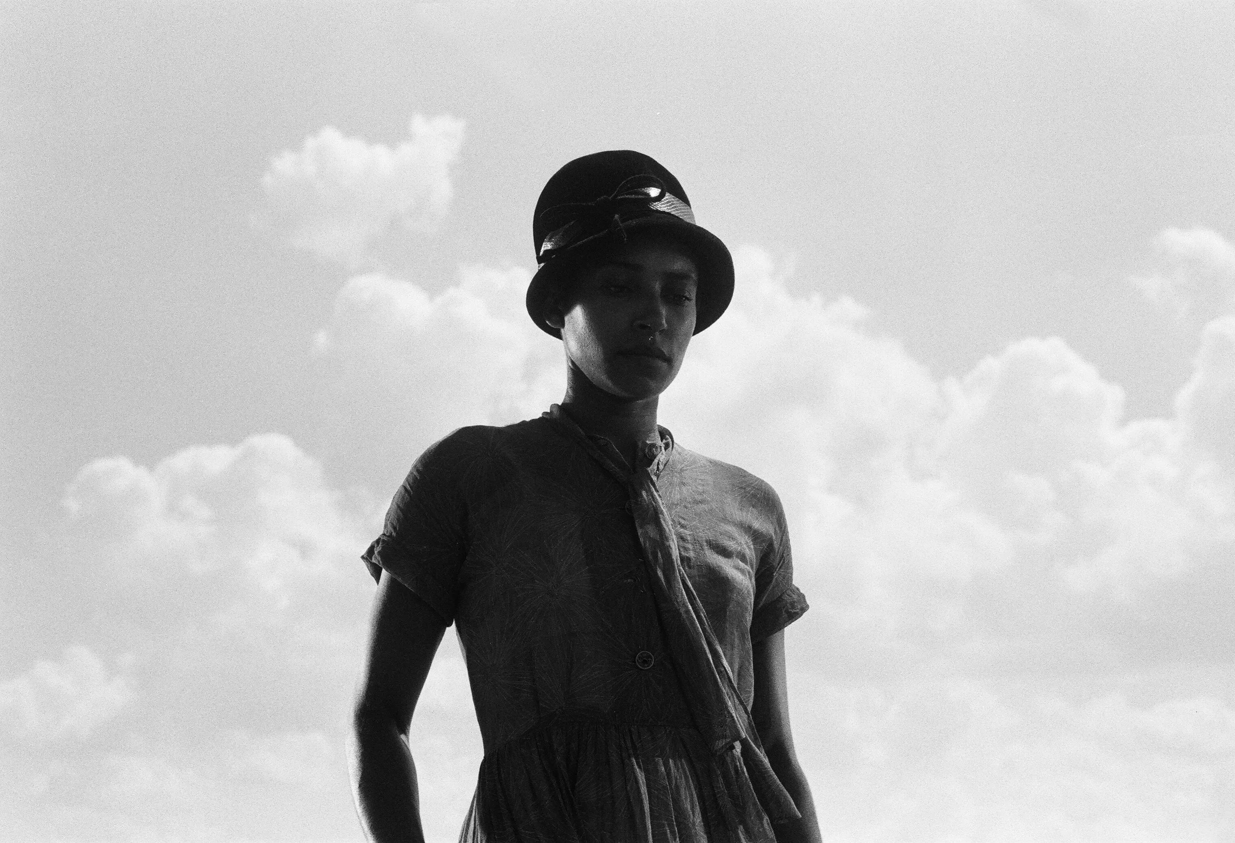 A woman wearing a hat and a short-sleeved shirt stands outdoors against a backdrop of a cloudy sky.