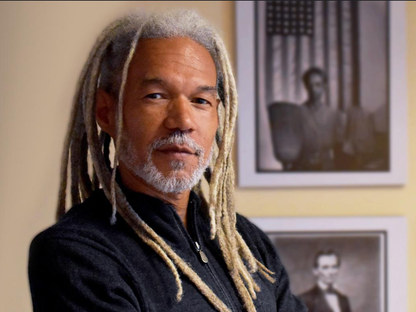 A man with gray dreadlocks and a gray beard standing indoors, with black and white photographs on the wall behind him.