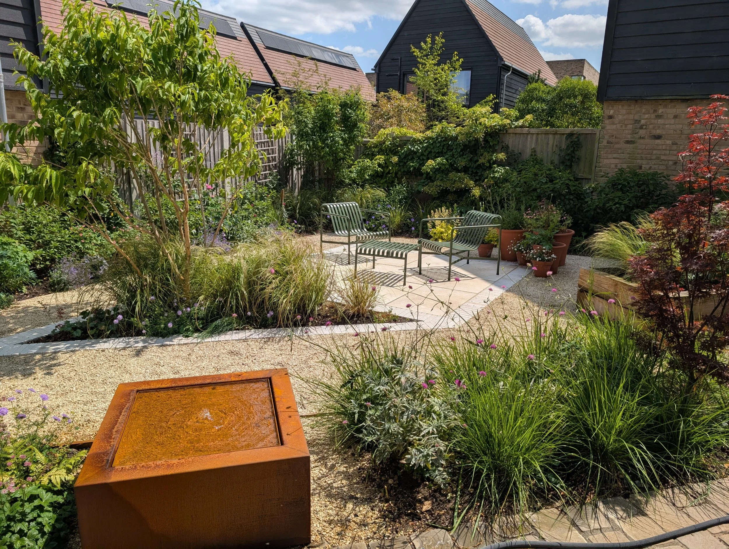 A landscaped garden with seating area, potted plants, trees, plants, and a corten steel water feature on a sunny day.