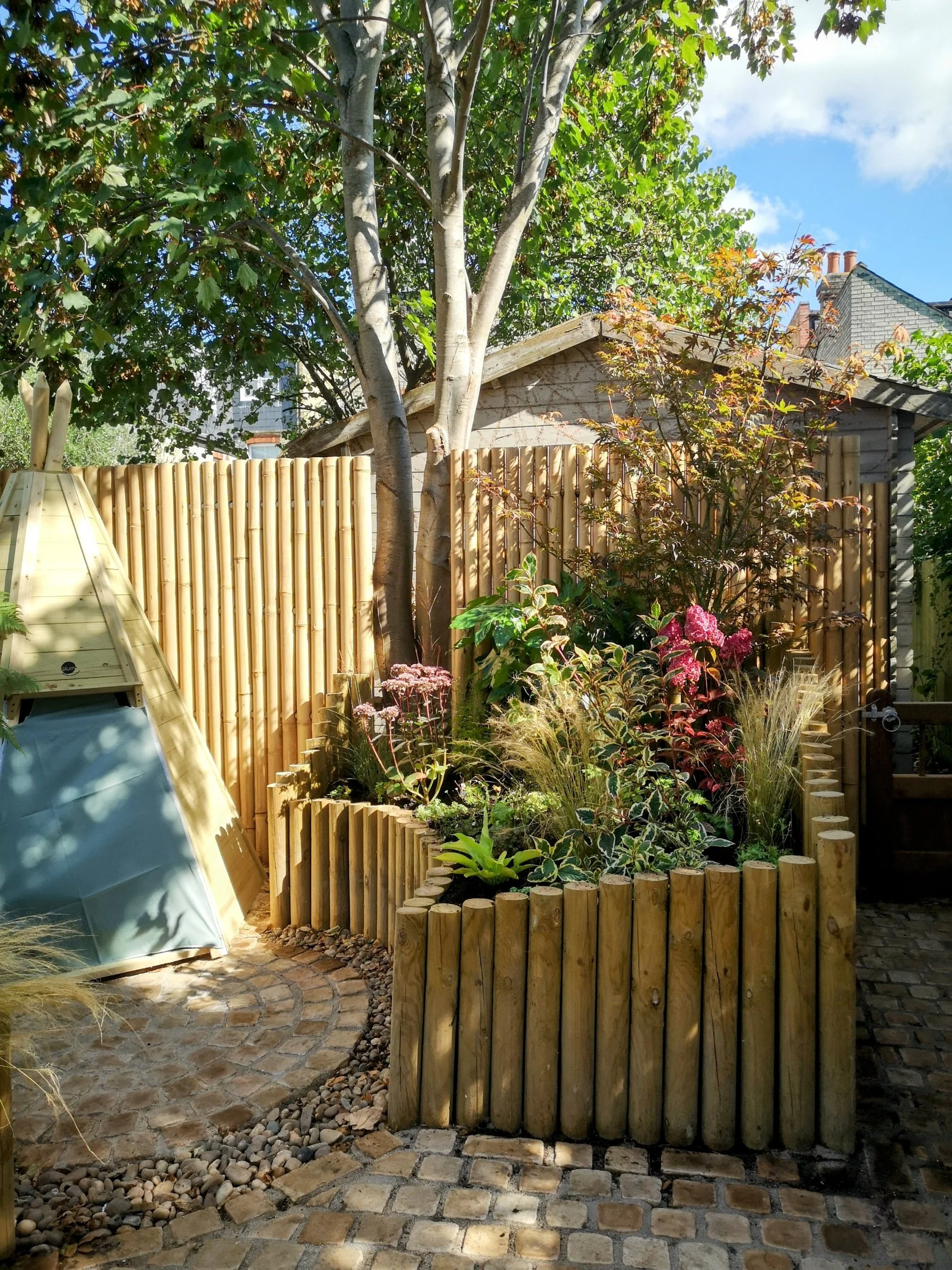 A backyard garden with a circular brick pathway, a raised flower bed edged with wooden logs, and various plants and trees, including a large tree with a light-colored trunk and green leaves, under a blue sky with some clouds.