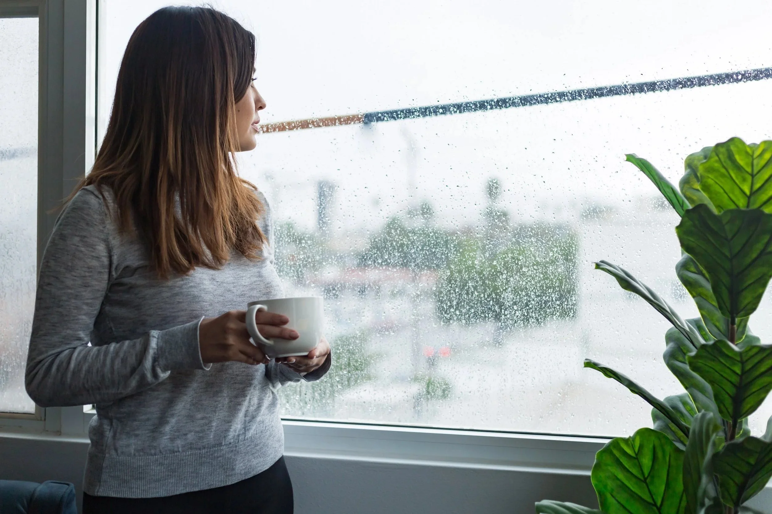 Person standing by a window on a rainy day, holding a mug and looking outside.
