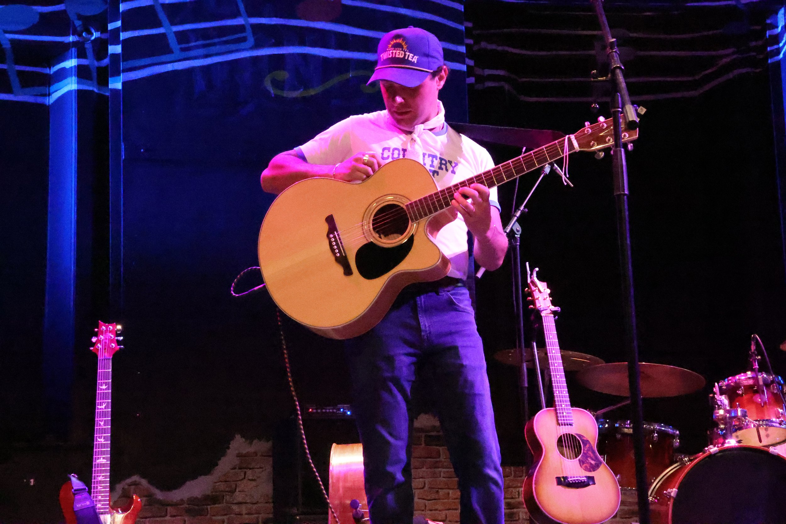 A man wearing a Twisted Tea cap and a white shirt with a scarf is playing an acoustic guitar on a stage illuminated with purple lighting. Several other guitars and musical equipment are visible in the background.