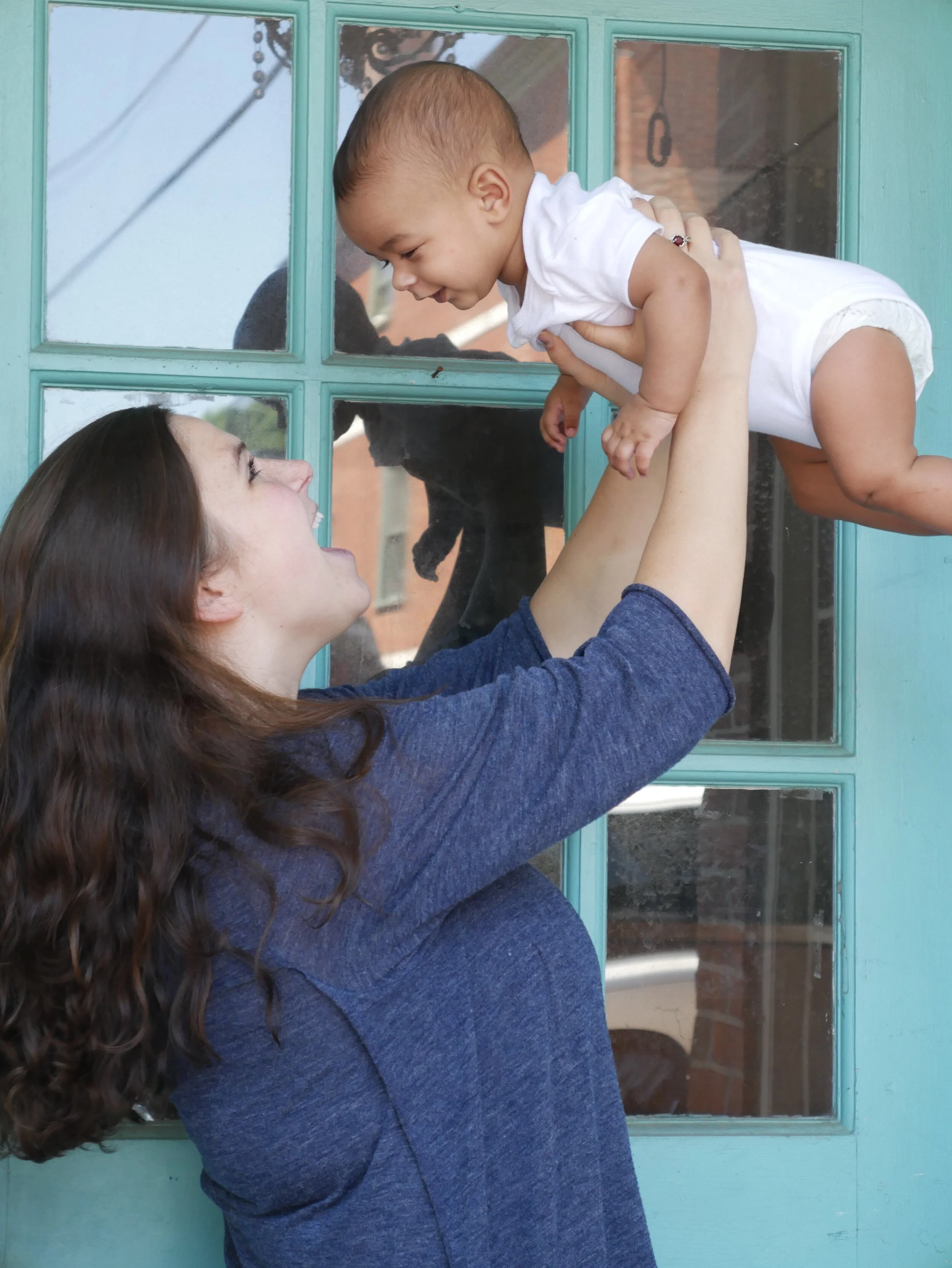 A young woman with long brown hair and wearing a dark blue shirt lifts a smiling toddler boy with short hair through a glass window in a turquoise door. The child is dressed in a white shirt and shorts, and they are engaging in a playful moment outdo