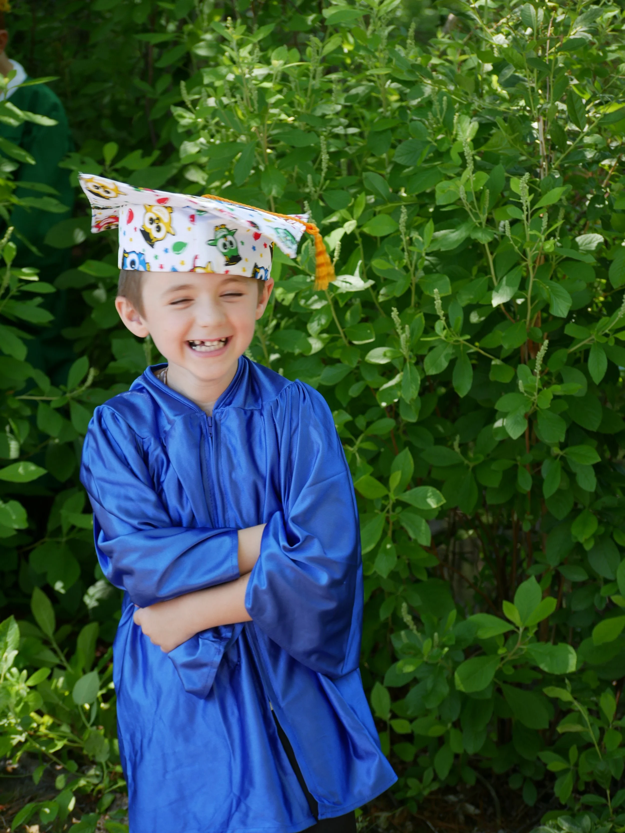 A young boy smiling with his eyes closed, wearing a blue rain jacket and a colorful graduation cap with cartoon characters, standing in front of green bushes.