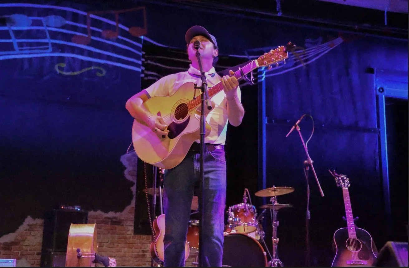 A man wearing a cap and glasses, singing into a microphone while playing an acoustic guitar on a stage with musical instruments and dark background.
