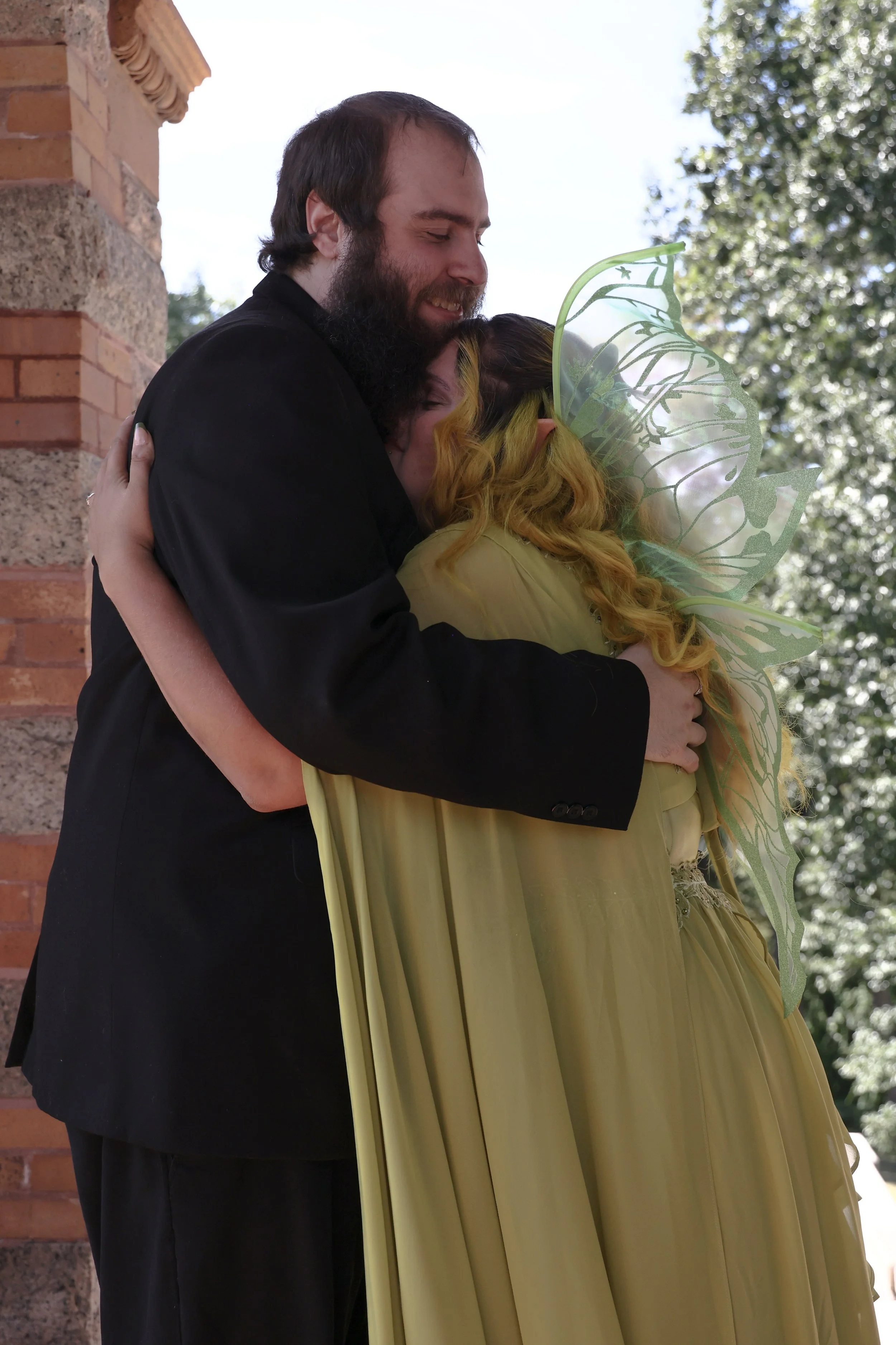 A man with a beard and a woman with yellow curly hair are hugging outdoors, with the woman holding a large, decorative green and silver leaf-patterned bouquet. They are standing next to a brick wall, and there are trees in the background.