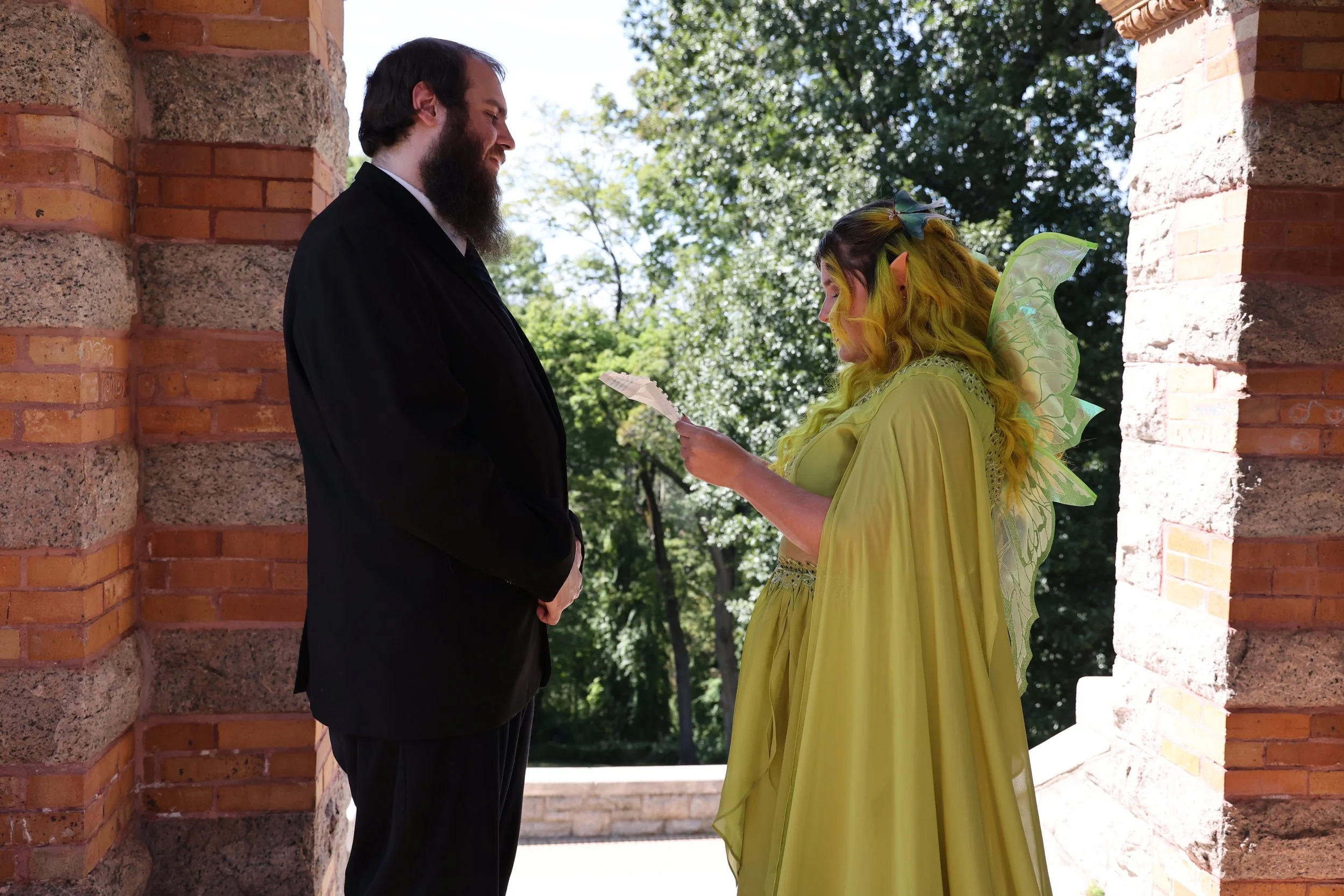 A man in a black tuxedo and a woman dressed as a fairy or fantasy character with yellow hair and wings are standing under a brick arch, with sunlight filtering through green trees in the background.