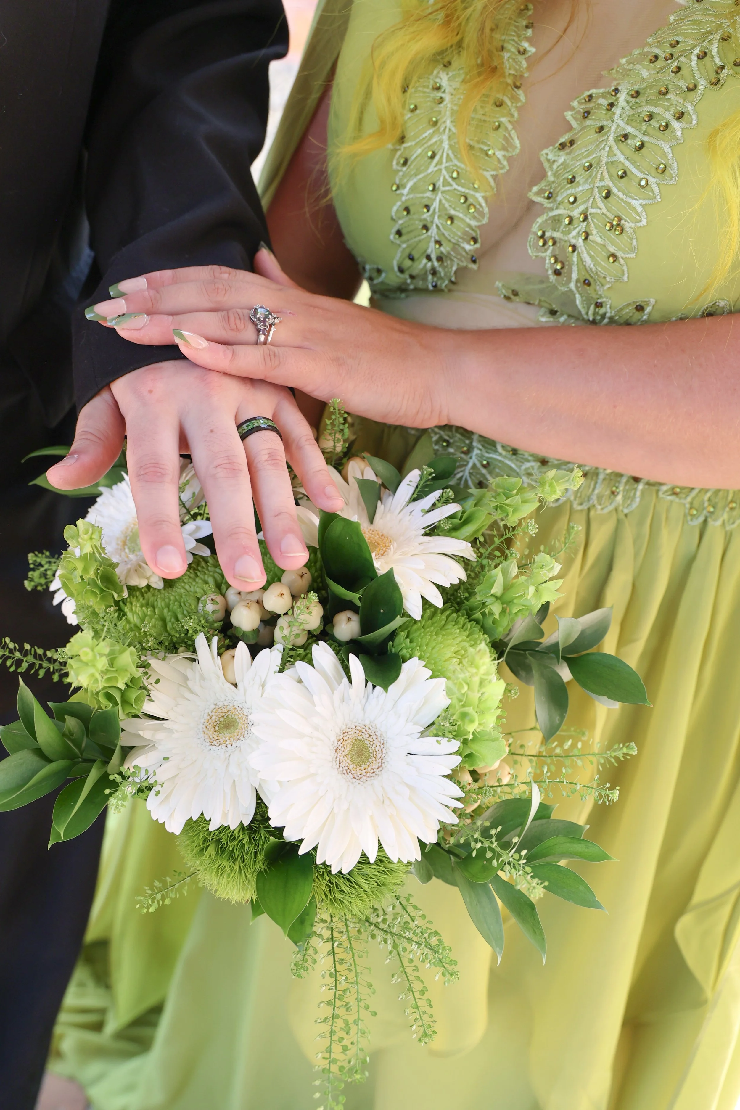 Close-up of a bride holding a bouquet of white and green flowers, with a woman wearing a bright yellow dress with embroidery and beads, and a man wearing a black suit. The bride's hand with a wedding ring is resting on the woman's hand, and the scene