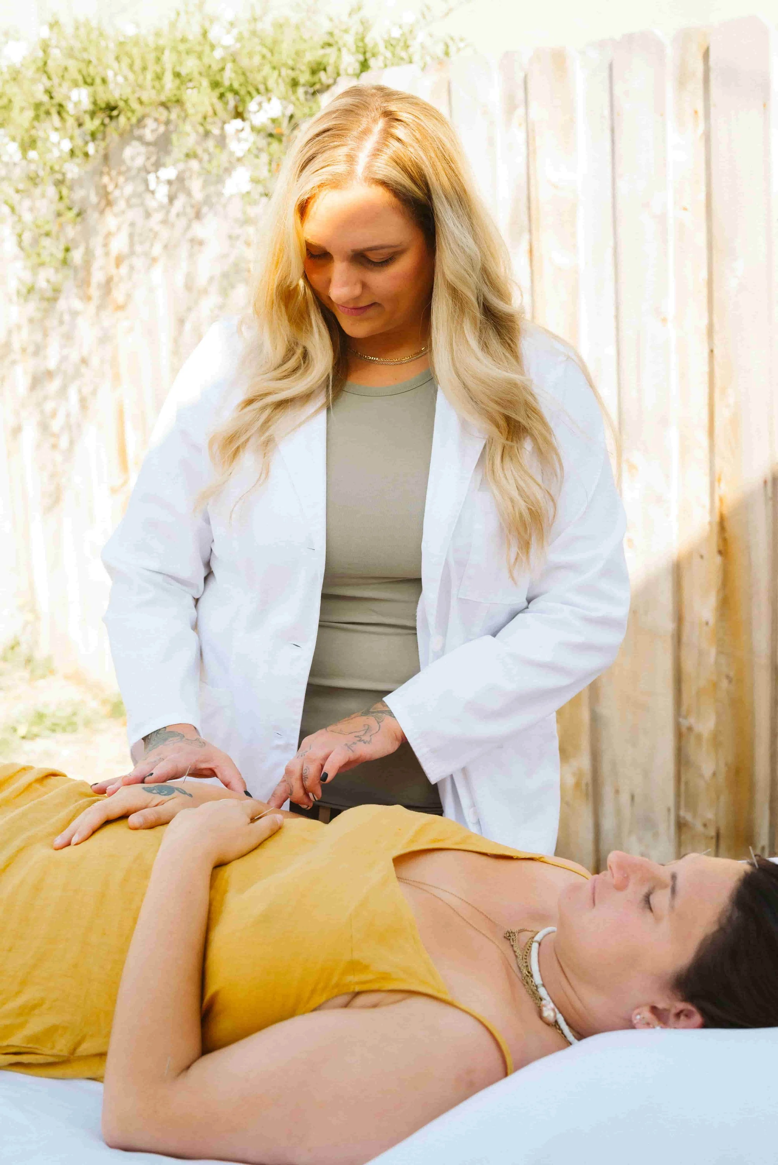 A woman in a yellow dress lying on a massage table while a therapist performs a massage. The woman has dark hair, wears jewelry, and appears relaxed. The background includes a wooden fence and outdoor foliage.