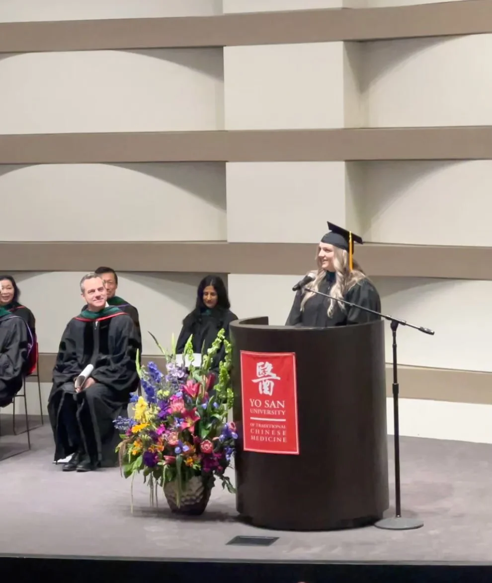 A graduation ceremony at Yo San University of Traditional Chinese Medicine, with students in academic regalia, one woman speaking at a podium decorated with a colorful flower arrangement.