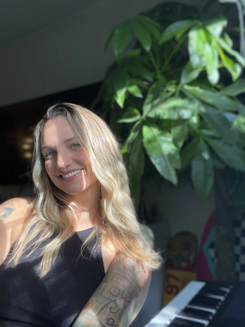 A woman with blonde hair taking a selfie, smiling, in a room with a large green leafy plant and a piano keyboard in the background.