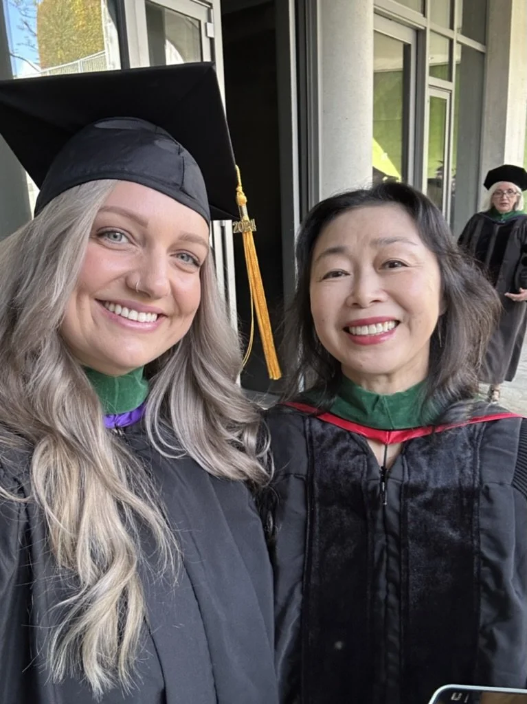Two women in academic regalia taking a selfie outdoors at graduation. One woman is wearing a graduation cap with a tassel and a gown, and the other is also in a gown, smiling.