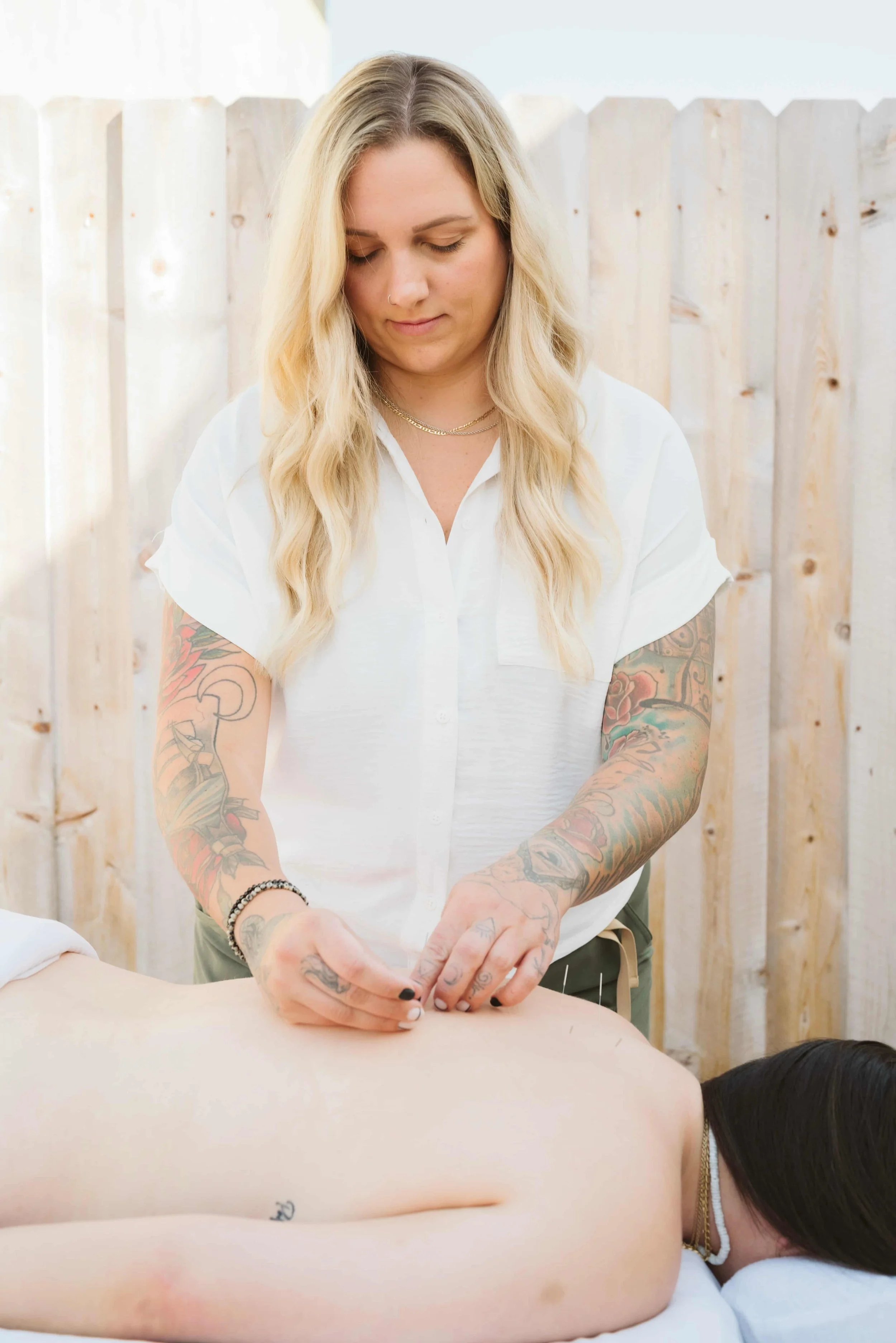 A woman with blonde hair and tattoos giving a massage or treatment to a person lying on a table, with a wooden fence in the background.
